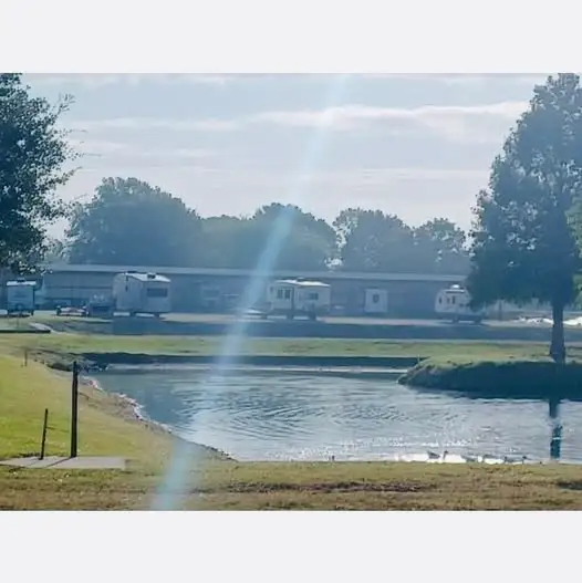Photo of a grassy pond with ducks in foreground and travel trailers parked beside a long building among trees.