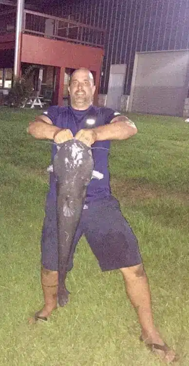 Man standing barefoot on grass at night holding up a very large catfish, with park buildings and picnic table behind.