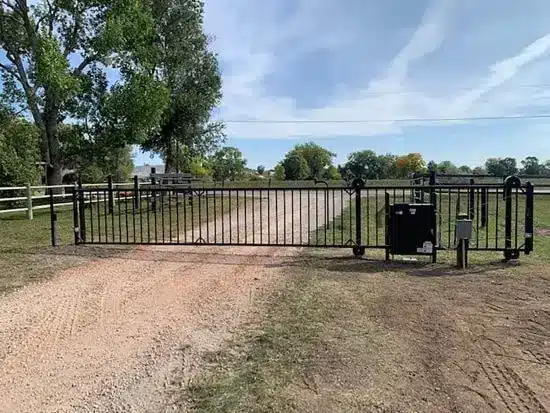 Black metal automated entrance gate with control box beside a gravel driveway, white fencing and large trees under blue